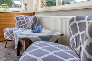 Un rincón amoblado con tapicería estampada, una mesa cubierta con un mantel azul y un ventanal con vistas a follaje.