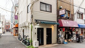 Une rue étroite en ville, bordée de bâtiments des deux côtés, avec un vélo garé sur le trottoir et une petite boutique dotée d’un auvent rouge.