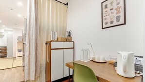 Une petite pièce avec une table en bois, une chaise verte, une bouilloire blanche et un tableau encadré accroché au mur.