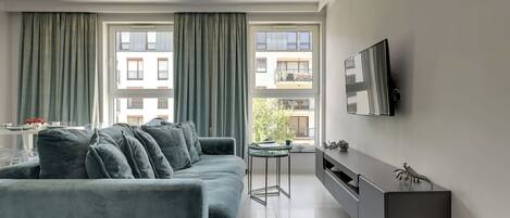 A modern living room with a grey sofa, a glass coffee table, a TV mounted on the wall, and a view of the outside through large windows.