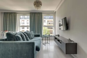 A modern living room with a grey sofa, a glass coffee table, a TV mounted on the wall, and a view of the outside through large windows. - Angielska Grobla 138B Atlantis | Near Old Town (Gdańsk)