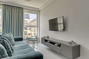 A modern living room with a grey sofa, a glass coffee table, a TV mounted on the wall, and a view of a residential area through the window. - Angielska Grobla 138B Atlantis | Near Old Town (Gdańsk)
