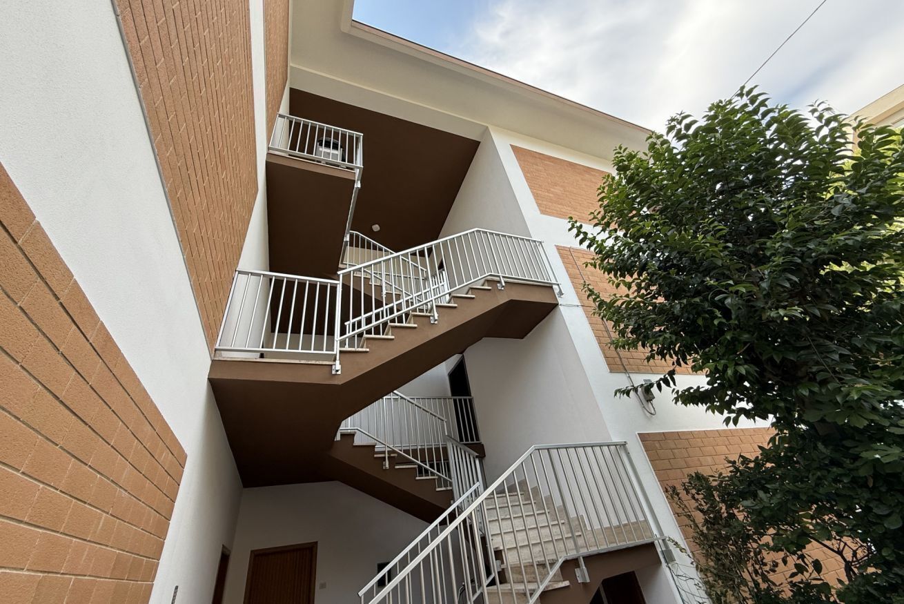 A multi-story residential building with a central staircase, balconies, and a tree in the foreground.