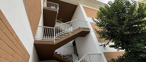 A multi-story residential building with a central staircase, balconies, and a tree in the foreground.