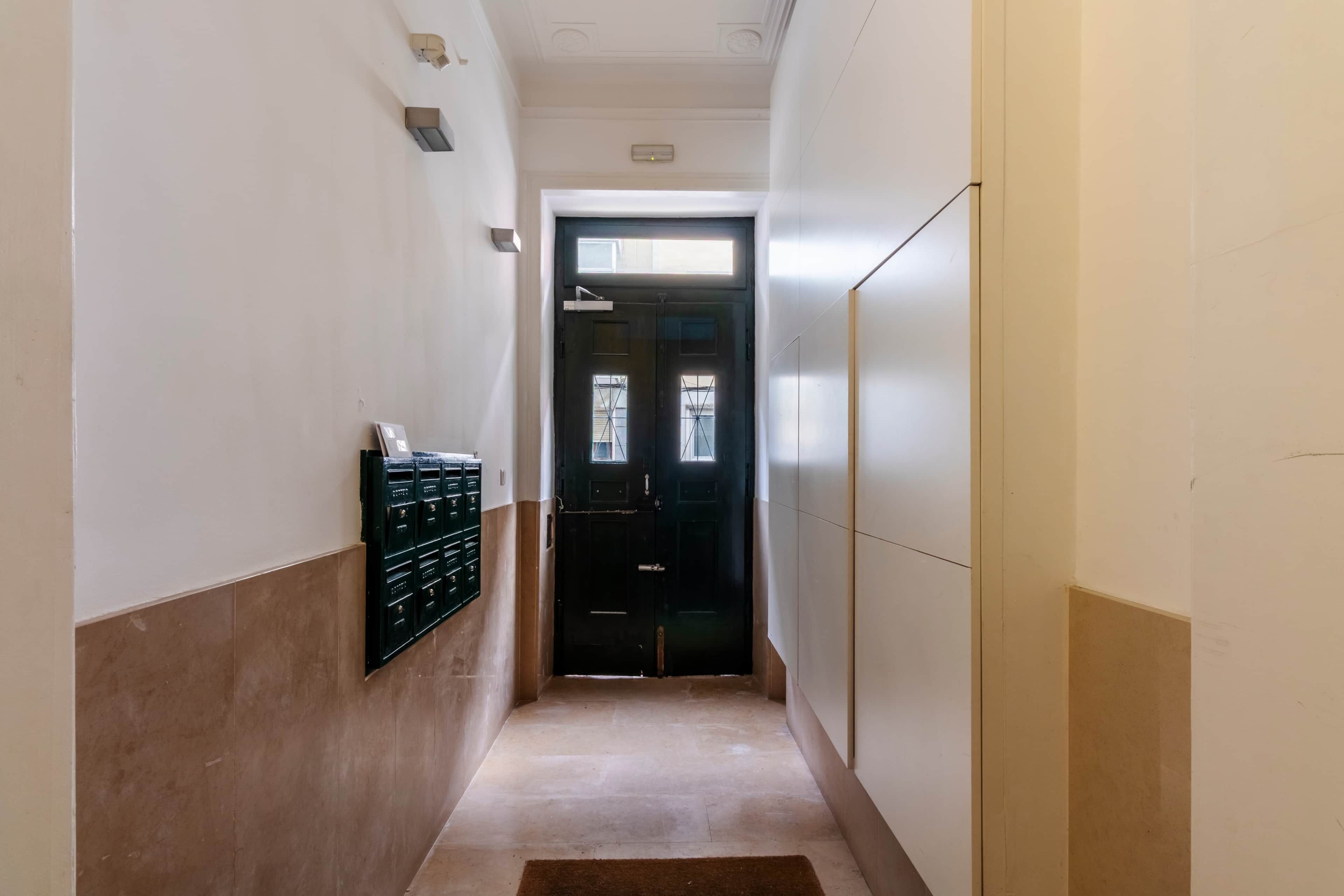 A narrow hallway with a black door at the end, a light switch on the wall, and a small window above the door.