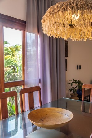 A dining area with a glass table, wooden chairs, a woven light fixture, and a view of greenery through a window. - Guesthouse Flamingo with 2 bedrooms and garden in the heart of Cumbuco (Caucaia)