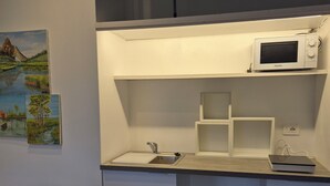 Une petite cuisine équipée d’un four à micro-ondes, d’un évier et d’un placard. Un tableau orne le mur et une chaise se trouve au premier plan.