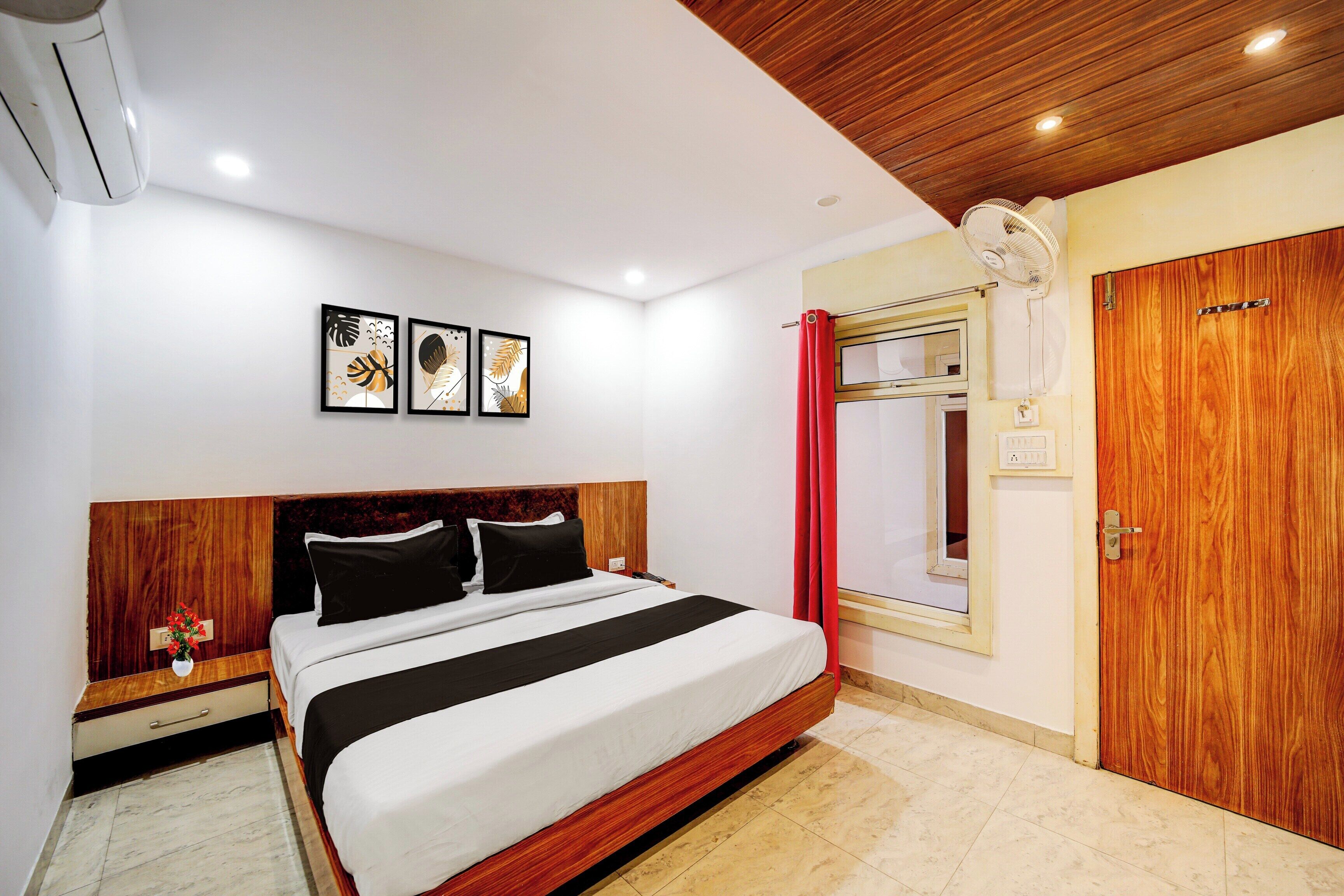 A hotel room with a bed, a wooden headboard, a bedside table with a vase, a red curtain, and a wooden wardrobe.