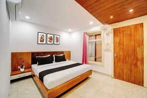 A bedroom with a wooden bed, white walls, and a wooden ceiling. There are three framed pictures on the wall, a window with pink curtains, and a wooden door. - Rani Kamlapati Railway Station (Bhopal)