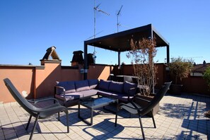 Eine Terrasse im Freien mit einer Eckcouch, zwei Liegestühlen, einem Couchtisch mit Glastischplatte und einer Pergola.