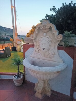 A stone fountain with a decorative head, surrounded by potted plants and a seating area. - Villa ROSALIA with a Jacuzzi, Barbecue, and Sunset View (Tonnara di Bonagia)
