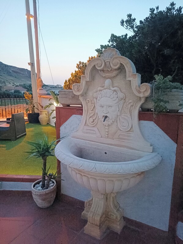 Ein Steinkopfbrunnen mit dekorativer Darstellung, umgeben von Blumentöpfen und einer Sitzgelegenheit.