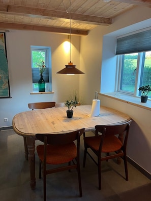 A dining area with a wooden table, four chairs, a hanging pendant light, and two windows with plants.