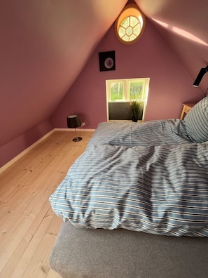 A bedroom with a sloped ceiling, a bed with striped bedding, a small table, a lamp, and a window with a view of greenery.