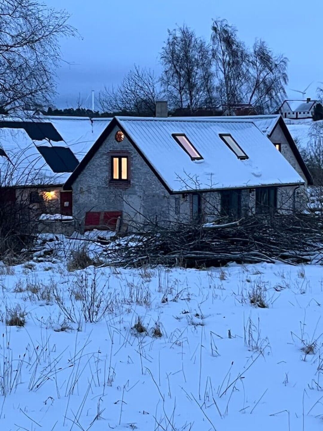 Una casa de piedra con techo inclinado y tragaluces, rodeada de nieve y árboles desprovistos de hojas.