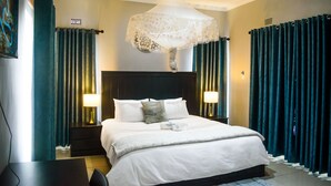 Hotellihuone, jossa on suuri sänky, kaksi yöpöytävalaisinta, sängyn päällä oleva koristeellinen kangas ja tummat verhot.
