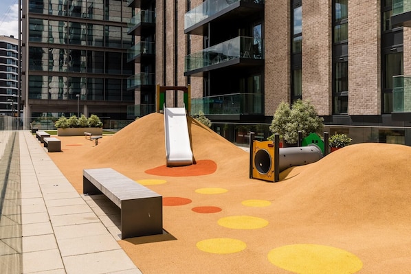 An outdoor play area with a slide, sand play area, and seating. - Landsby (Wembley)