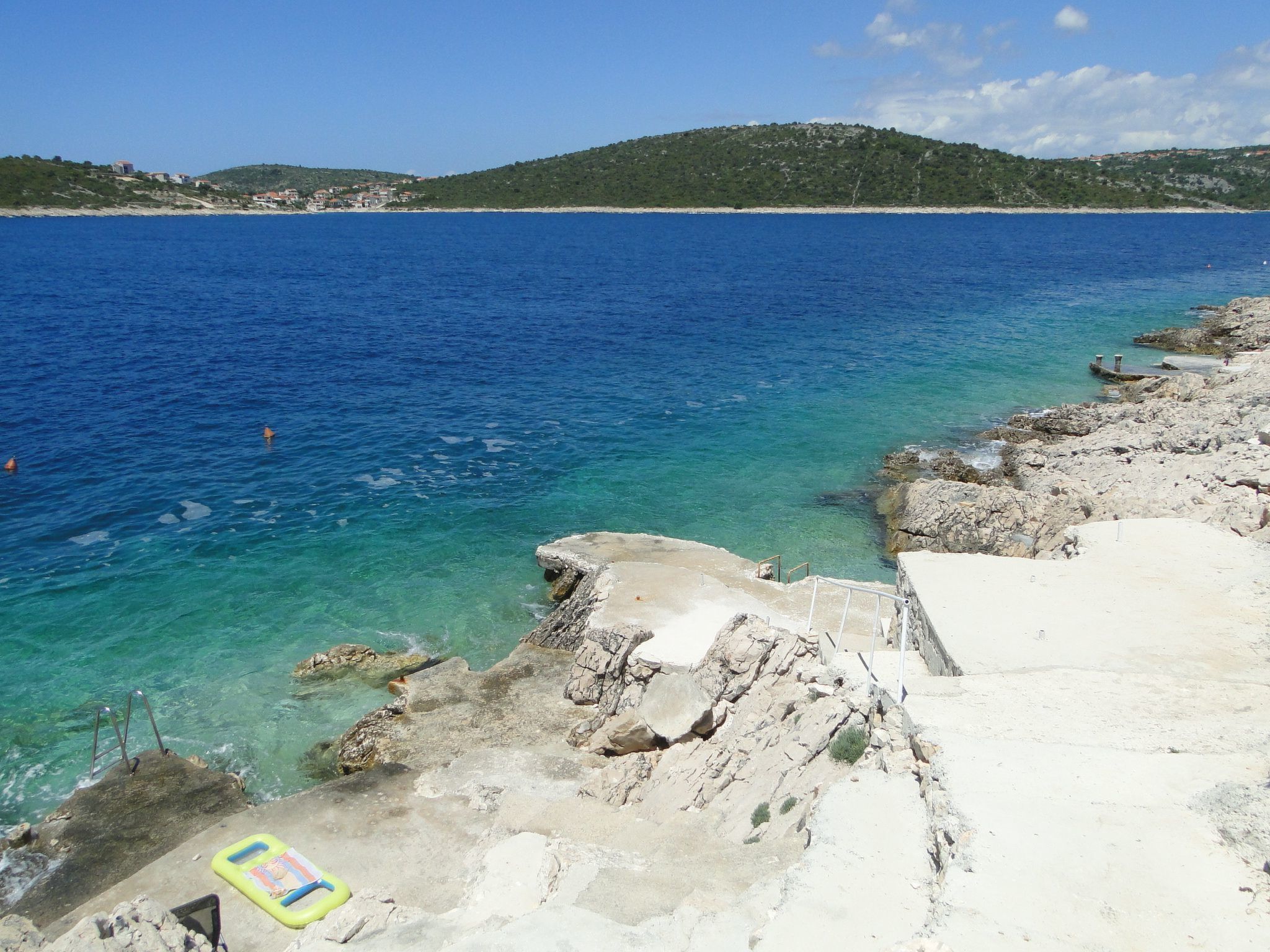 Un paysage côtier avec une eau d’un bleu limpide, une côte rocheuse et, au loin, une colline parsemée de bâtiments.