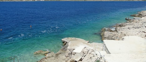 Un paysage côtier avec une eau d’un bleu limpide, une côte rocheuse et, au loin, une colline parsemée de bâtiments.