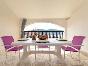 Un balcón con una mesa lista para dos personas, sillas, un florero con flores y una vista de un paisaje lejano.