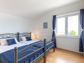 Ein Doppelbett mit Metallgestell und zwei Kissen, ein Nachttisch mit Lampe, ein Fenster mit Vorhängen und ein Wandbild.