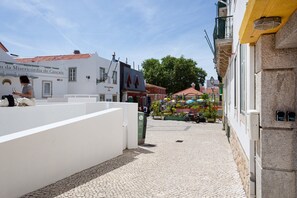 Eine Kopfsteinpflasterstraße mit Gebäuden, ein Café mit Sonnenschirmen und eine Person, die auf einer weißen Mauer sitzt.