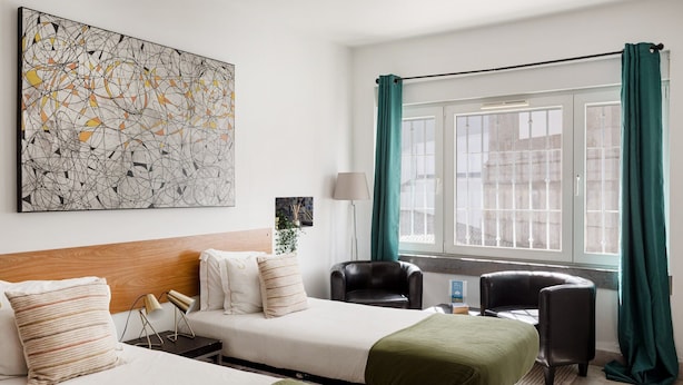 Una habitación de hotel con dos camas, un cuadro abstracto de gran tamaño, una ventana con cortinas, un escritorio con una lámpara y una silla.