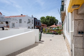 A cobblestone street with buildings, a café with umbrellas, and a person sitting on a white wall. - Twin Studio Steps From Praia da Rainha, Cascais (Cascais)