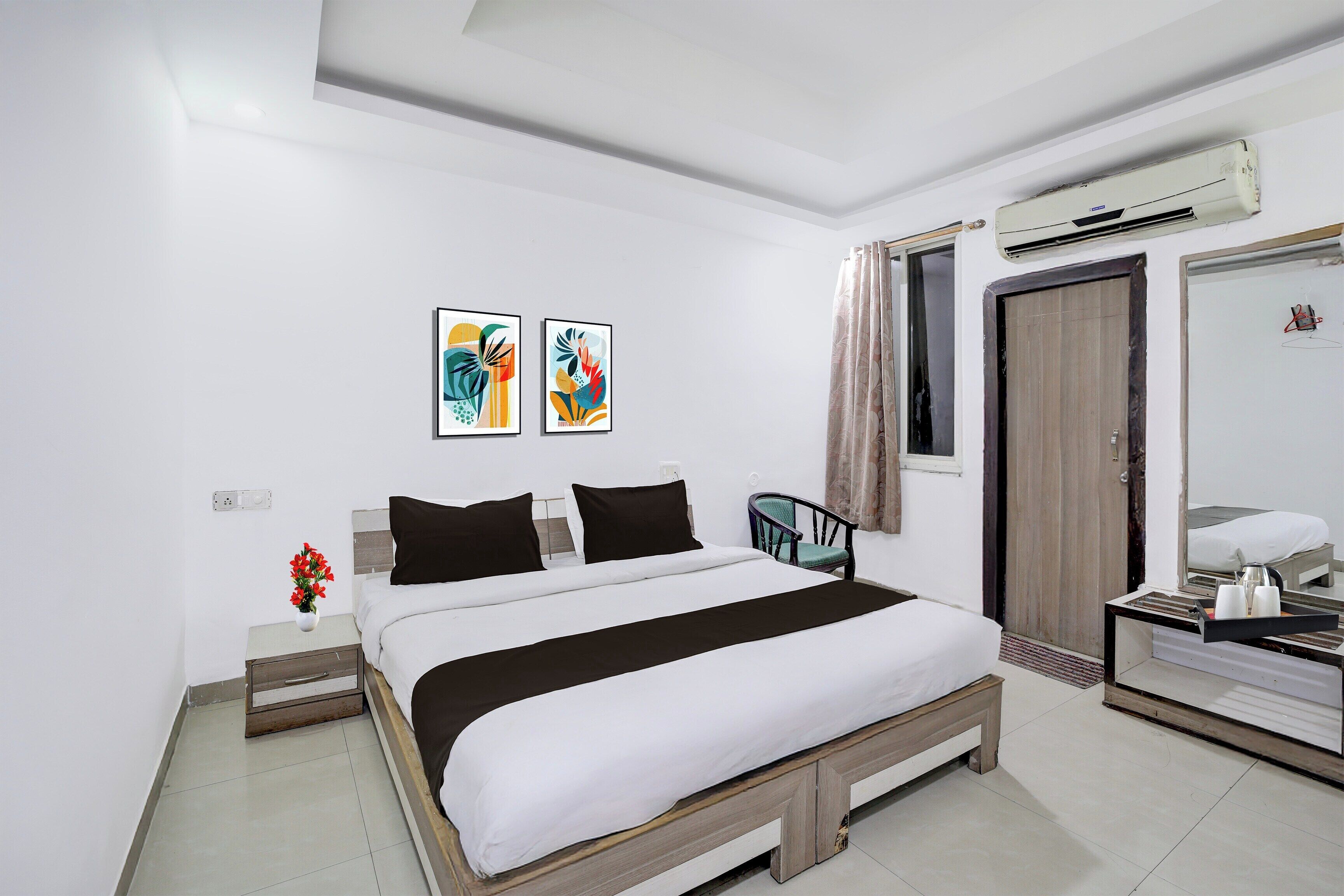 A hotel room with a bed, a chair, a desk, an air conditioner, and two artworks on the wall.