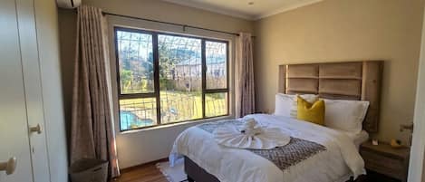A bedroom with a large window, a bed with white bedding, a wooden floor, and a headboard.