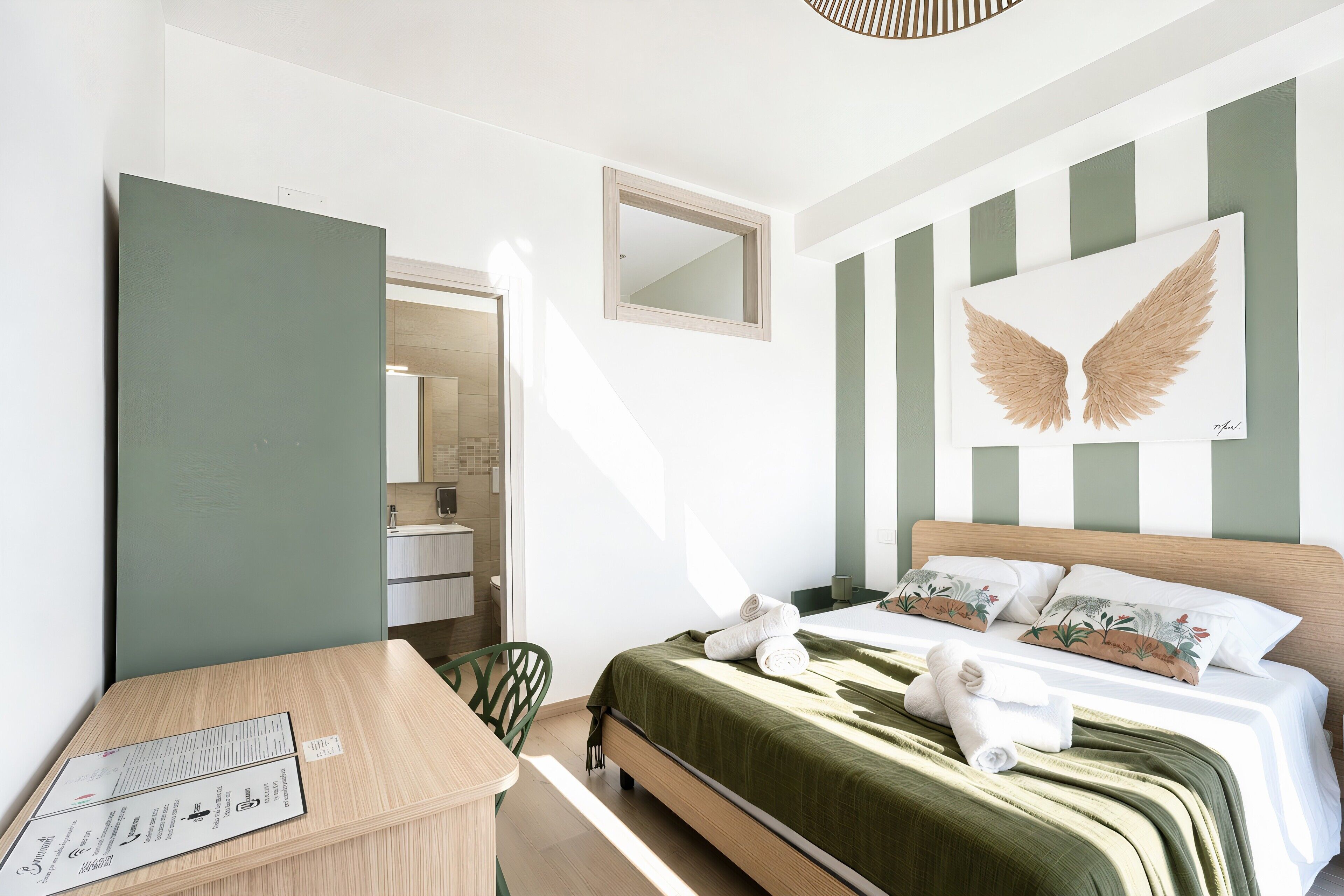 A hotel room with a bed, a desk, a chair, a mirror, and a wall with a large winged artwork.