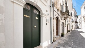 Una calle estrecha con un edificio que presenta una puerta doble verde oscuro, fachada blanca y un pequeño balcón con plantas.