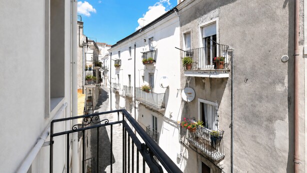 A narrow street with buildings on either side, balconies with potted plants, and a clear blue sky. - ‘Domus Dell’Angelo 4’ – Comfort and Authenticity in the Heart of Monte Sant’Angelo (Monte Sant'Angelo)
