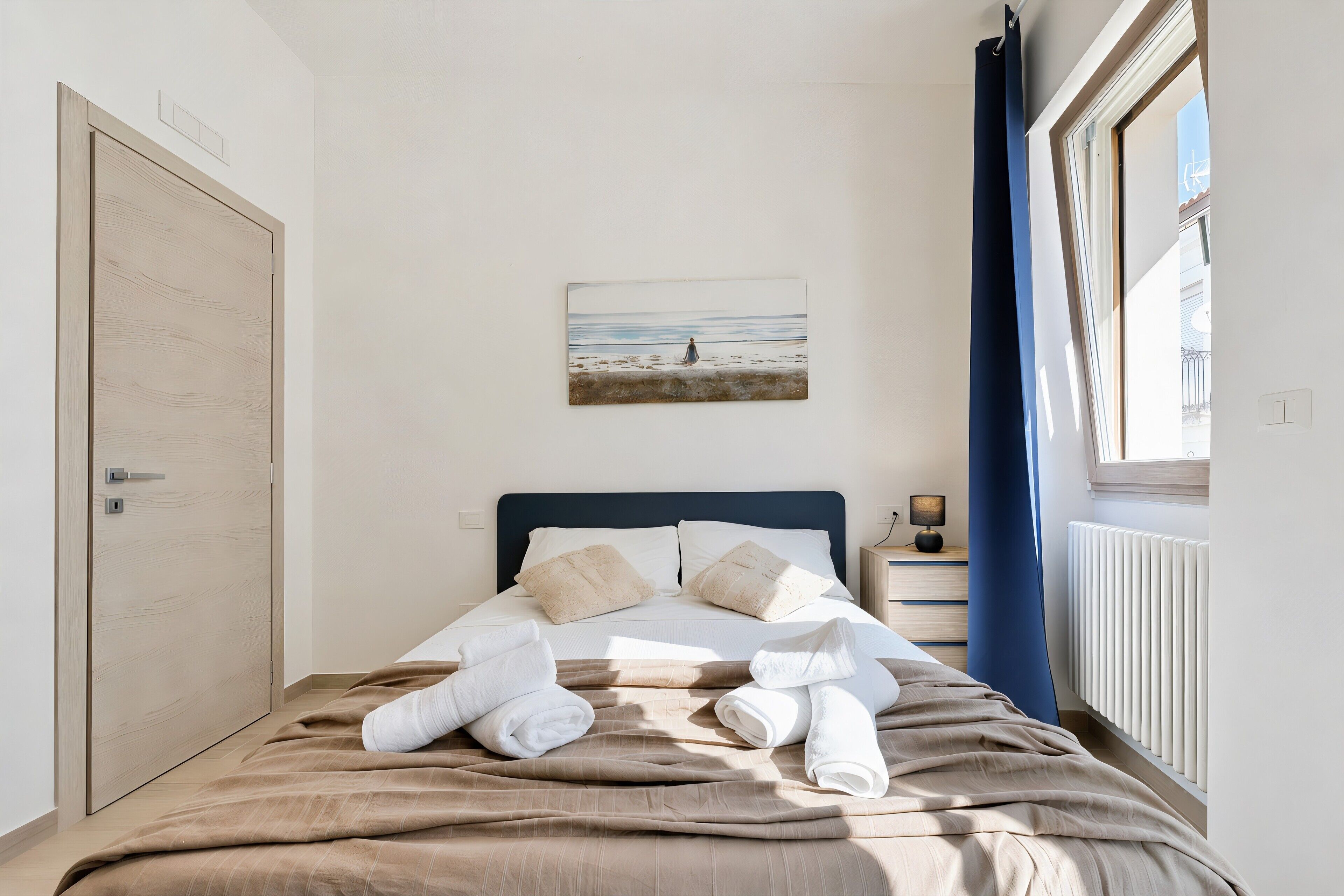 Una cama bien hecha con toallas, un cuadro de una playa y una ventana con cortinas.