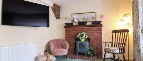 Una sala de estar con televisión de pantalla plana, chimenea de ladrillo, un sillón rosa, una silla de madera y un otomano estampado.