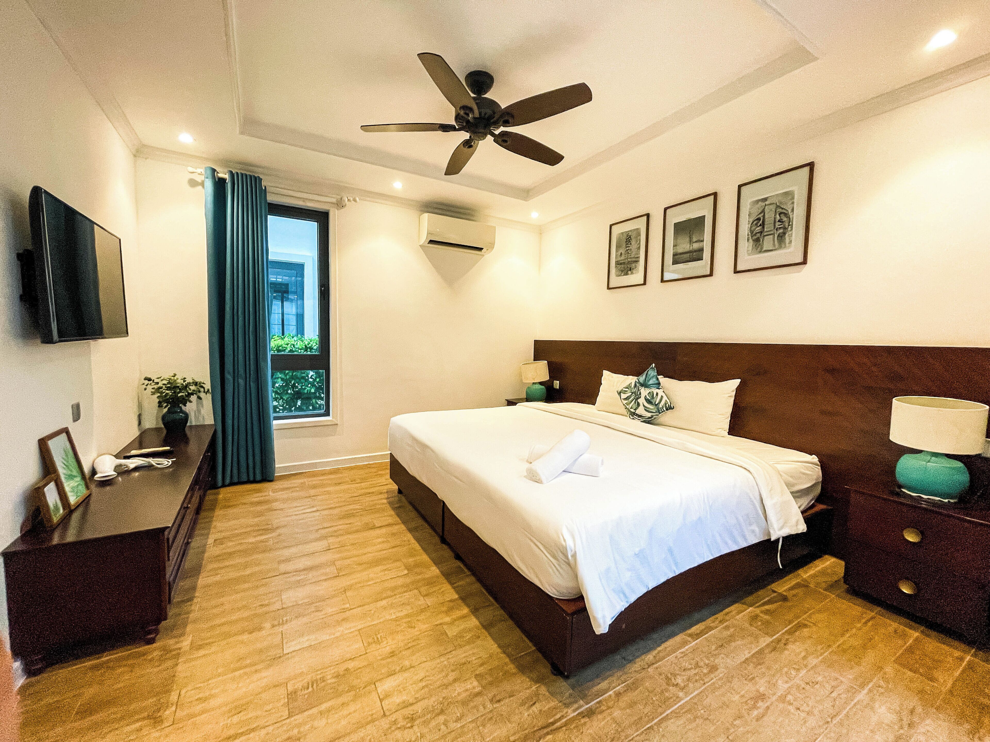 A hotel room with a bed, a ceiling fan, a TV, a window with curtains, and framed pictures on the wall.