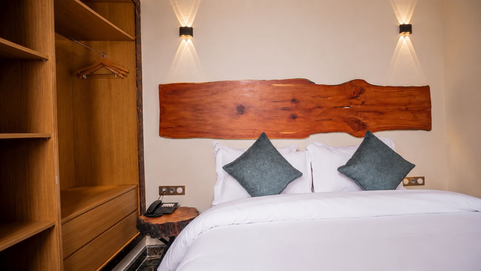 A hotel room with a wooden headboard, a bed with white linens, two pillows, a bedside table with a phone, and a wooden wardrobe.