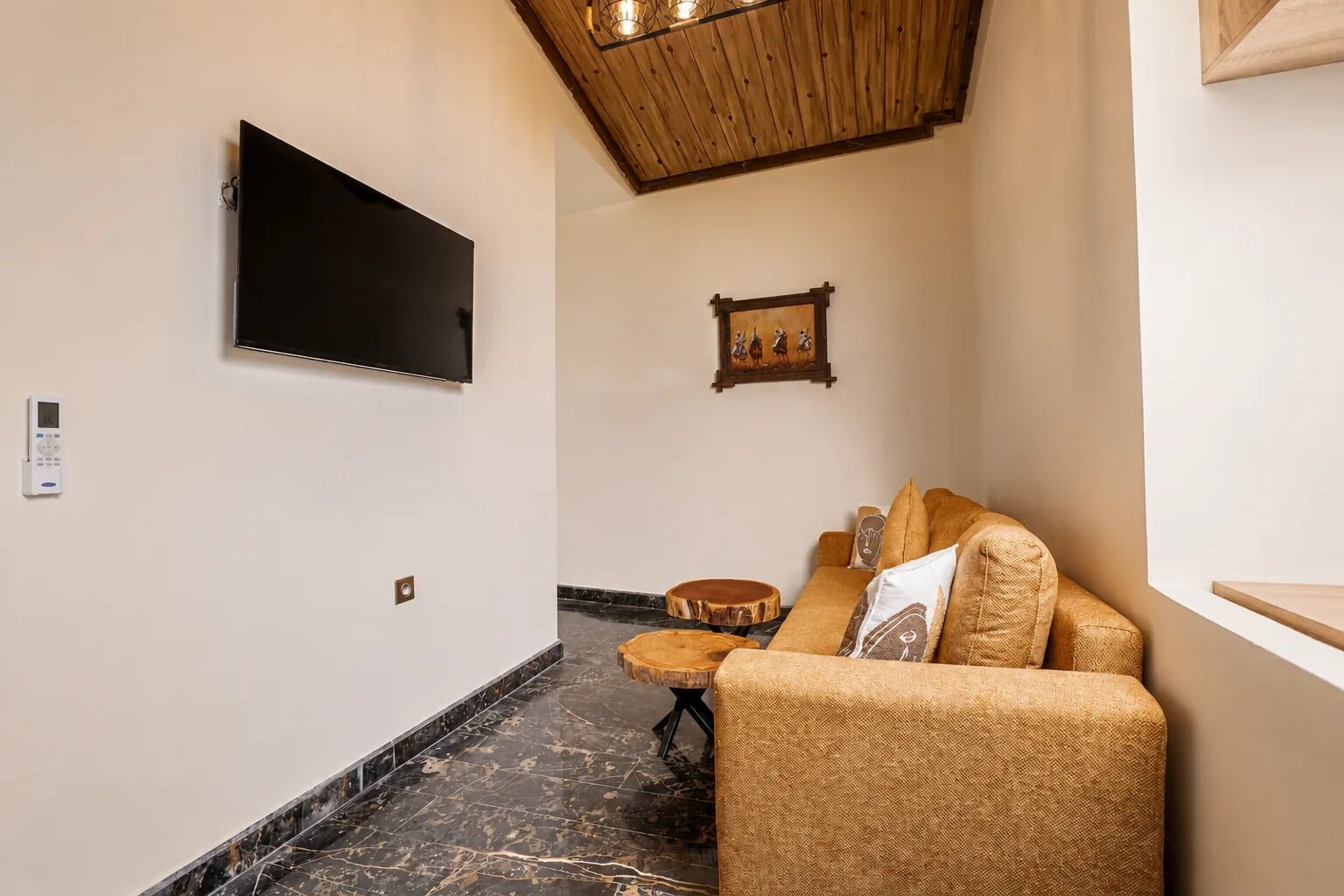 Un pasillo con un televisor de pantalla plana montado en la pared, techo de madera, un cuadro enmarcado, un sofá de mimbre con cojines y una pequeña mesa redonda.