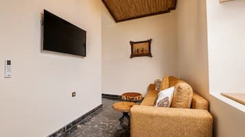 Un pasillo con un televisor de pantalla plana montado en la pared, techo de madera, un cuadro enmarcado, un sofá de mimbre con cojines y una pequeña mesa redonda.