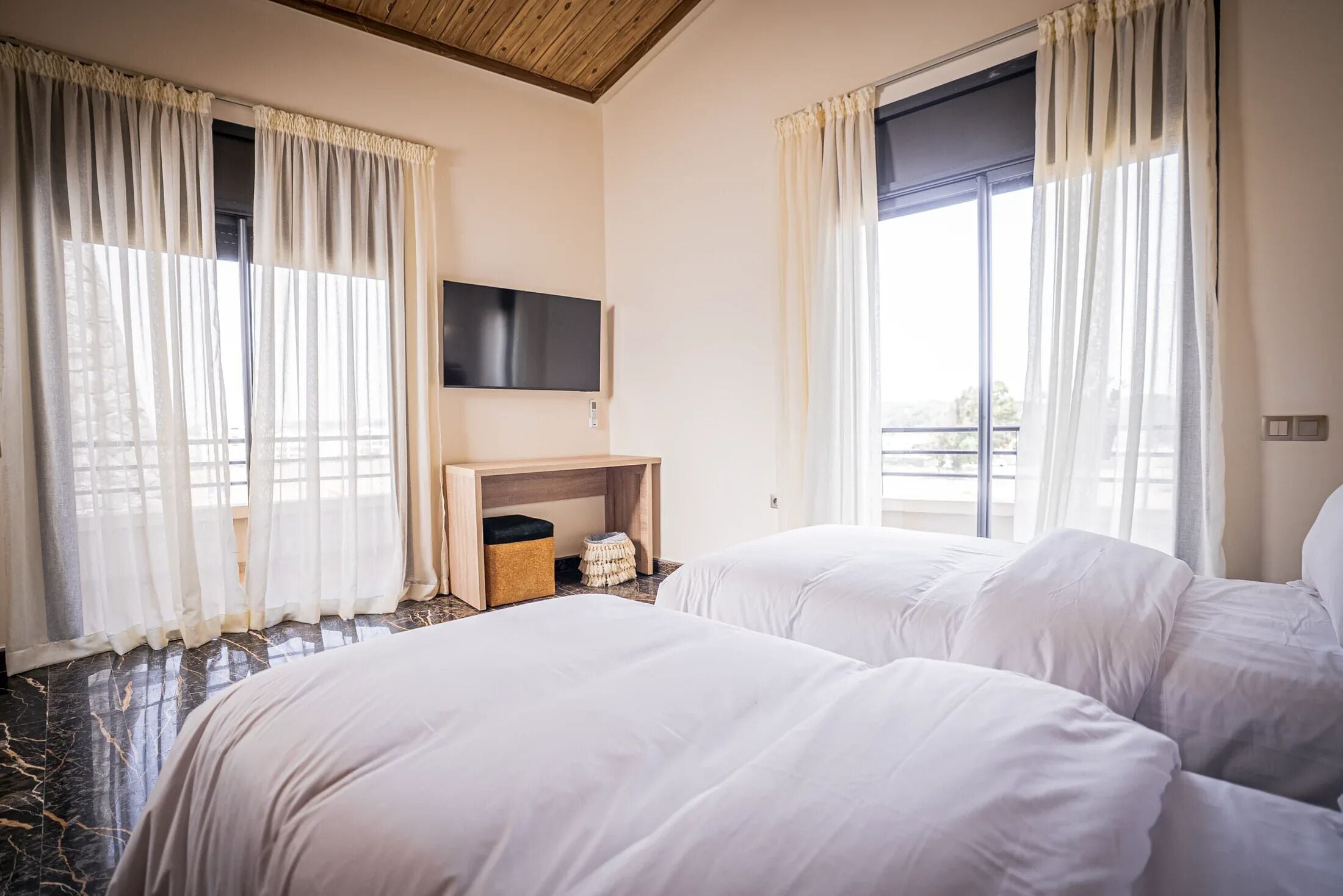 Une chambre d’hôtel avec deux lits, un téléviseur à écran plat fixé au mur, une petite table d’appoint en bois et une fenêtre avec des rideaux légers.