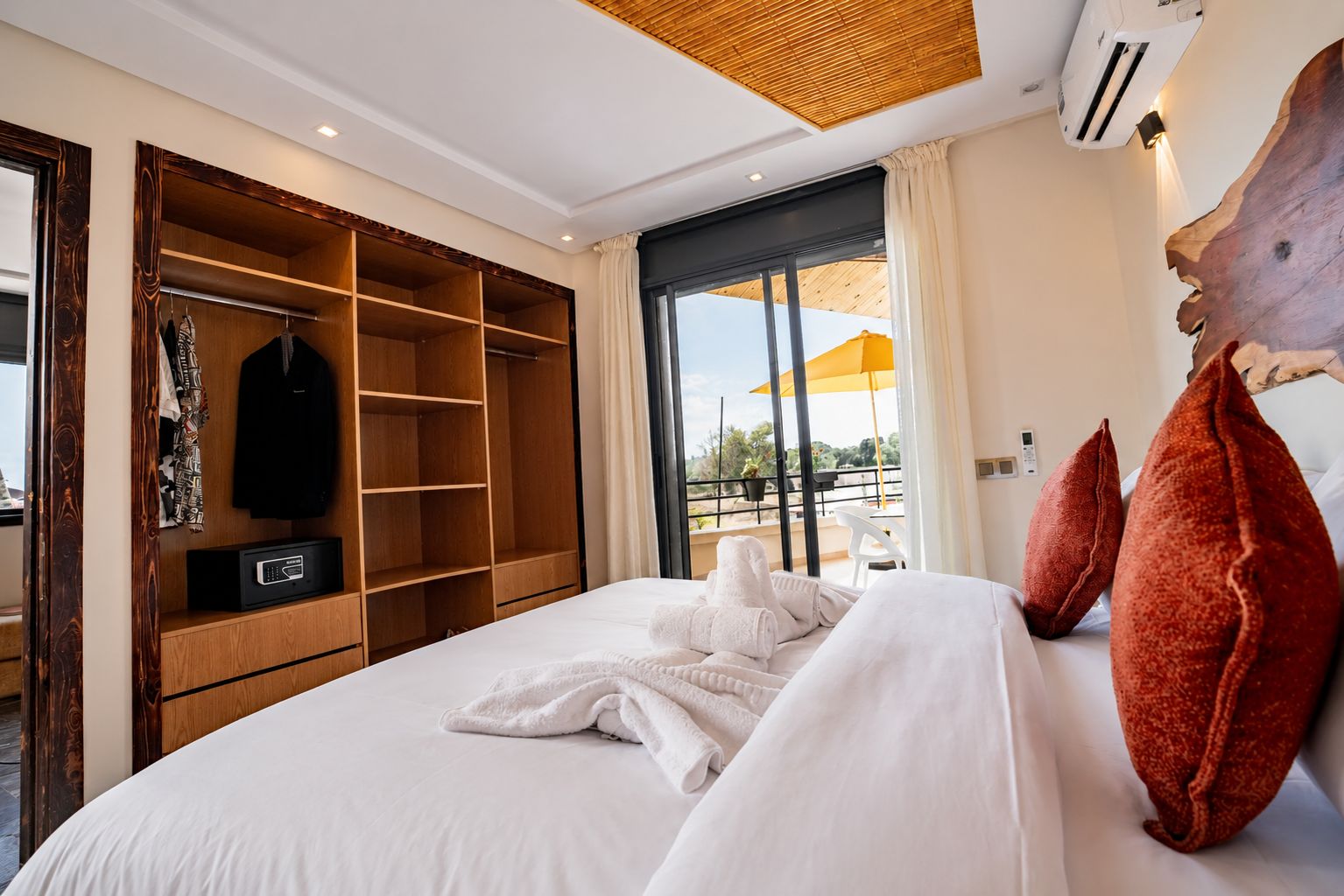 A hotel room with a large bed, a wooden wardrobe, a balcony with a yellow umbrella, and a large wooden headboard.
