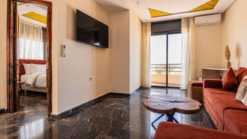 A hotel room with a bed, a flat-screen TV, a wooden coffee table, a red sofa, and a balcony with curtains.