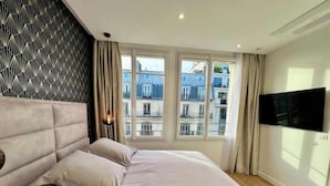 A bedroom with a large bed, a TV mounted on the wall, and a view of buildings through the window. - Very bright, recently renovated apartment with air conditioning (Île-de-France)