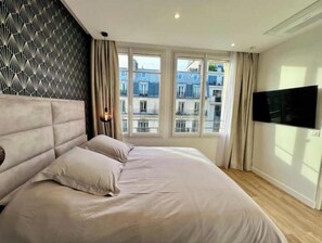 Ein Schlafzimmer mit einem großen Bett, einem an der Wand befestigten Fernseher und Blick auf Gebäude durch das Fenster.
