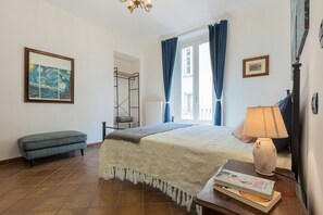 Ein Schlafzimmer mit einem Bett, einer Bank, einem Nachttisch mit Lampe und einem Fenster mit Vorhängen.