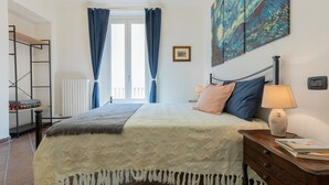 Um quarto com cama, mesa de cabeceira, abajur, um quadro e uma janela com cortinas.