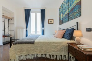 Ein Schlafzimmer mit Bett, Nachttisch, Lampe, einem Gemälde und einem Fenster mit Vorhängen.