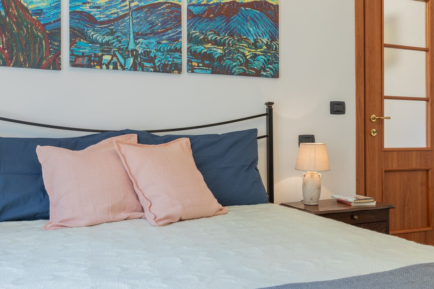 Un dormitorio con cama, almohadas, una mesita de noche con lámpara y decoración en la pared.