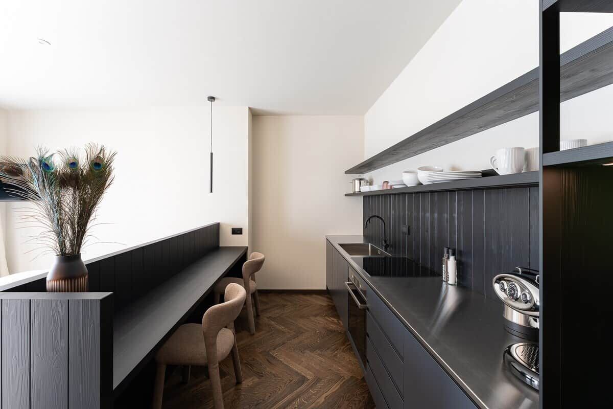 Une cuisine moderne avec un plan de travail foncé, un four encastré et un évier. On y trouve des tabourets de bar et une plante en pot.