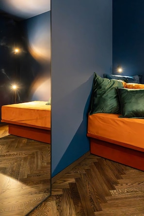 Une chambre moderne avec un canapé et un lit, tous deux dotés de linge de lit orange. La chambre a un plancher en bois et un mur d’accent bleu.